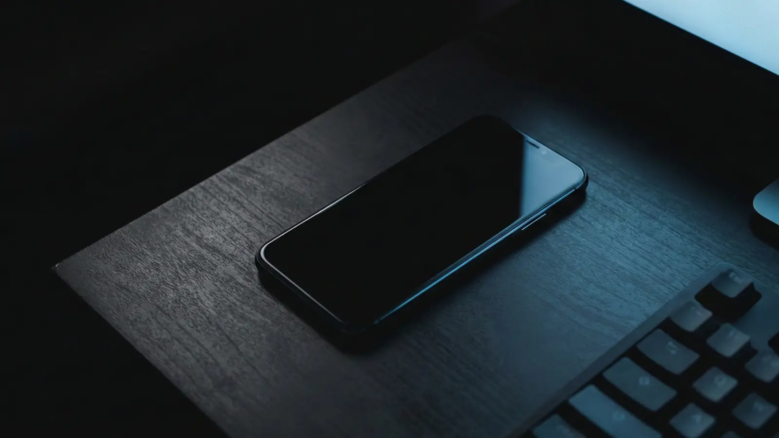 A top-down, atmospheric macro photograph in a dark room at night. A modern smartphone is placed face down on a dark, matte desk surface, symbolizing the removal of all outside distractions.